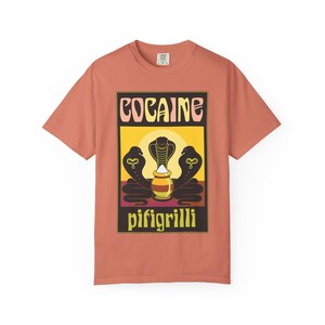 May include: A coral-coloured short-sleeve t-shirt featuring a rectangular graphic print. The design displays the text COCAINE at the top and pifigrilli at the bottom, surrounding three black cobra silhouettes and a central pot with white powder.