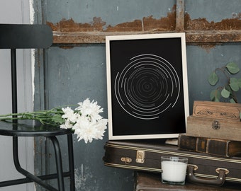May include: A framed black and white abstract art print featuring concentric circles. The artwork is displayed on a vintage suitcase, alongside a candle, wooden boxes, and white flowers. A black stool and a distressed blue wall complete the scene.