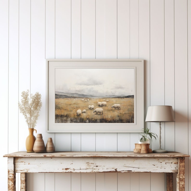 May include: A framed landscape print depicts a field of sheep grazing under a cloudy sky. The artwork features a muted color palette of beige, brown, and gray, creating a serene and pastoral scene. The print is displayed on a rustic wooden table with decorative vases and a lamp.
