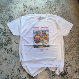 May include: White t-shirt featuring a graphic of a boxing match between Hearns vs Leonard. The graphic includes the text "WORLD WELTERWEIGHT CHAMPIONSHIP" and "CAESARS PALACE". The shirt is made of a soft material.