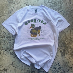 Puede incluir: Camiseta blanca con un gráfico de pato y el texto "GORE-TEX fabrics". El pato está en un estanque con lluvia. El texto de abajo dice: "Para aquellos que no entrarán por la lluvia". Una camiseta informal de manga corta.