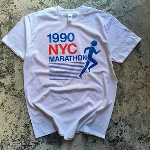May include: White t-shirt with the text "1990 NYC MARATHON" in red and blue. A blue silhouette of a runner is also on the shirt. The shirt is made of a soft material and has short sleeves.