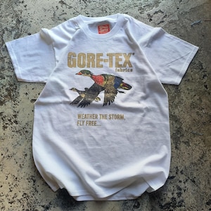 Puede incluir: Camiseta blanca con el logo dorado "GORE-TEX fabrics" y una ilustración de dos patos en vuelo. El texto "WEATHER THE STORM, FLY FREE..." está impreso debajo. La camiseta tiene una etiqueta roja.