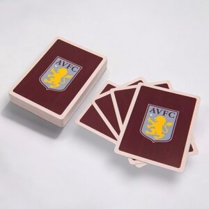 May include: A deck of playing cards with a maroon back and the AVFC logo. The logo features a yellow lion on a blue shield with the letters AVFC. The deck is stacked next to a few fanned-out cards.