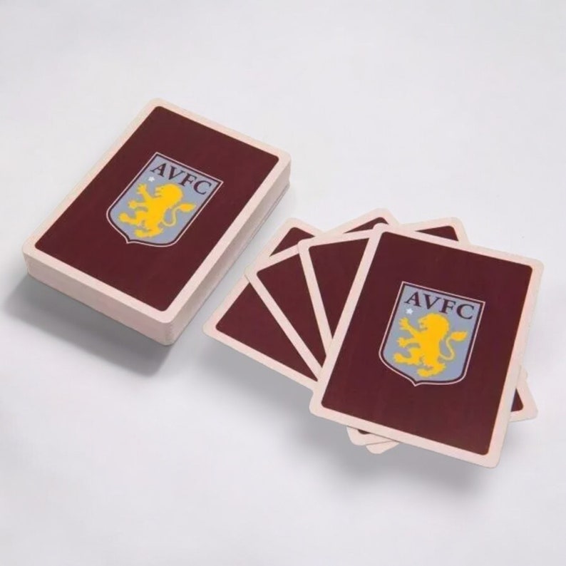 May include: A deck of playing cards with a maroon back and the AVFC logo. The logo features a yellow lion on a blue shield with the letters AVFC. The deck is stacked next to a few fanned-out cards.
