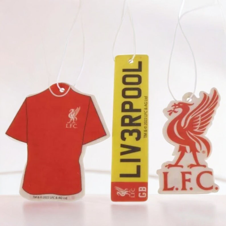 May include: Three car air fresheners featuring the Liverpool Football Club logo. The first is a red t-shirt with the LFC logo. The second is a yellow rectangle with the text "LIV3RPOOL" and the LFC logo. The third is a white bird with the LFC logo and the text "L.F.C."