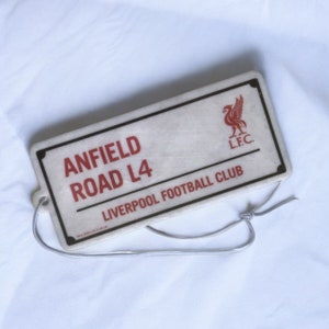 May include: White and red car air freshener with a black border. It features the text "Anfield Road L4 Liverpool Football Club" and a red Liverpool Football Club crest with the letters "L.F.C."