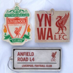 May include: Three car air fresheners featuring the Liverpool Football Club logo. The first air freshener is red, green, and white with the text "You'll Never Walk Alone" and "Liverpool Football Club". The second air freshener is white with red text that says "YNWA" and "L.F.C." The third air freshener is white with black text that says "Anfield Road L4" and "Liverpool Football Club".