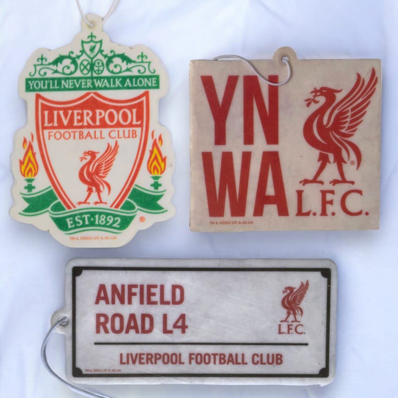 May include: Three car air fresheners featuring the Liverpool Football Club logo. The first air freshener is red, green, and white with the text "You'll Never Walk Alone" and "Liverpool Football Club". The second air freshener is white with red text that says "YNWA" and "L.F.C." The third air freshener is white with black text that says "Anfield Road L4" and "Liverpool Football Club".