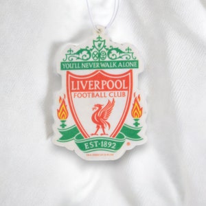 May include: A white car air freshener with the Liverpool Football Club crest. The crest is red, white, and green with a red bird and the words "You'll Never Walk Alone" and "Liverpool Football Club".