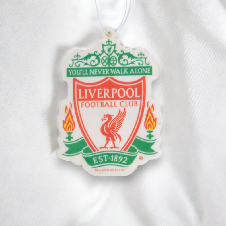 May include: A white car air freshener with the Liverpool Football Club crest. The crest is red, white, and green with a red bird and the words "You'll Never Walk Alone" and "Liverpool Football Club".