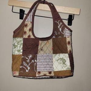 Puede incluir: Un bolso tote de patchwork con un diseño acolchado. El bolso presenta una variedad de telas marrones, beige y crema en un patrón geométrico. El bolso tiene un asa marrón y está suspendido de una percha de madera.