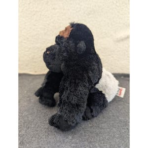 May include: A black plush gorilla toy with brown and white details. The gorilla is depicted in a seated posture, with its arms and legs extended. The toy has a soft, furry texture and a tag attached to its rear.