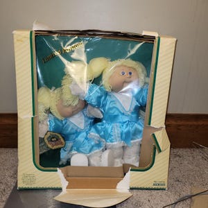 May include: Two Cabbage Patch Kids dolls in a box. The dolls have blonde yarn hair, blue eyes, and wear light blue satin dresses with white lace trim. The box is labelled "Limited Edition" and has a green interior.