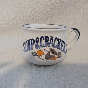 May include: White ceramic soup mug with a blue rim and handle. The mug features the words "SOUP & CRACKERS" in blue lettering, along with illustrations of vegetables. The handle is also blue.