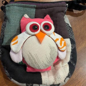 May include: A patchwork fabric pouch with a pink owl applique. The owl has large red and black eyes, an orange beak, and white and orange wings. The pouch is made of various fabrics in shades of gray, black, and brown.