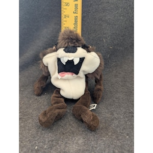 May include: A plush Taz, the Tasmanian Devil, toy. The plush toy has a white face, large white teeth, and a pink tongue. The fur is brown and the toy is approximately 7 inches tall.