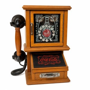 Puede incluir: Un teléfono Coca-Cola vintage de madera con un auricular y un dial negros. El teléfono tiene un armario con el logotipo de Coca-Cola y un cajón con un asa de metal. El teléfono es un artículo decorativo.