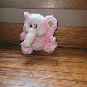 May include: A soft pink elephant toy with a white head and trunk. The elephant has large, round eyes and a tag attached to its ear. The toy is made of a soft, fuzzy material and is on a wooden surface.