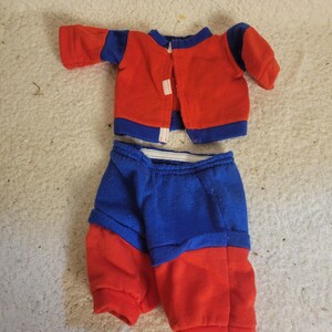 Könnte beinhalten: Ein zweiteiliges Puppenoutfit bestehend aus einer langärmeligen Jacke und einer passenden Hose. Die Jacke ist überwiegend rot mit blauen Ärmeln und einem blauen Kragen und hat eine Öffnung vorne. Die Hose ist an der Taille blau und an den Beinen rot.