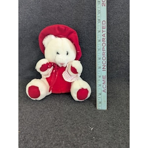 May include: A plush white teddy bear wearing a red bonnet and dress. The bear has red accents on its paws and ears. A red ribbon with a bow adorns the bear's neck. The bear is approximately 15 cm tall.