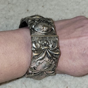 Può includere: Un bracciale a polsino argentato con dettagli in rilievo floreali e simili a insetti. L'ampio bracciale si avvolge attorno al polso, mostrando un design vintage dettagliato. La chiusura è visibile e il bracciale sembra essere in metallo.