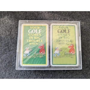 May include: A clear plastic case containing two playing cards. Each card has a green background with the text "YOUR GOLF GAME'S IN BIG TROUBLE WHEN..." and a cartoon illustration of a golfer and a red creature.