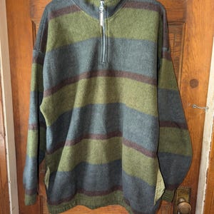 May include: A fleece pullover with horizontal stripes in shades of green, gray, and brown. The long-sleeved top has a quarter-zip closure and a relaxed fit. The garment is displayed against a wooden door.