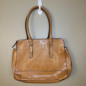 May include: A tan-colored handbag with two handles and a rectangular shape. The bag has a glossy finish and features metal hardware on the handles and a zipper closure. The bag is hanging from a white hook.