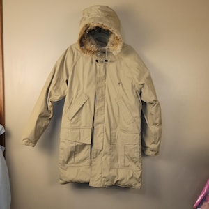 May include: Tan parka with a hood trimmed with faux fur. The coat has a button and zipper closure, multiple pockets, and a small logo on the chest. The parka is designed for cold weather and outdoor activities.