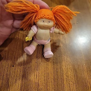 Puede incluir: Una muñeca vintage de Cabbage Patch Kids con cabello de hilo naranja peinado en coletas. La muñeca lleva un atuendo rosa y calcetines rosas. Sostiene un objeto amarillo en su mano derecha.