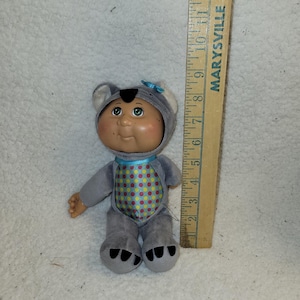 May include: A Cabbage Patch Kids doll wearing a gray koala bear costume with a blue bow. The doll has a round face, rosy cheeks, and green eyes. The costume has a colorful polka-dot pattern on the front. A wooden ruler is next to the doll.