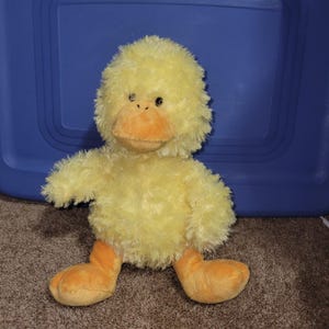 Puede incluir: Un juguete de peluche de pato amarillo con una textura esponjosa. El pato tiene un pico y patas naranjas, y un pequeño ojo negro. El juguete está sobre una superficie marrón con un fondo azul.