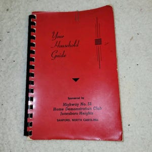 May include: A red booklet titled "Your Household Guide" with black spiral binding. The cover features stylized text and is sponsored by the Highway No. 53 Home Demonstration Club in Sanford, North Carolina. A silver paper clip secures the pages.