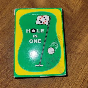 May include: A vintage deck of playing cards with a golf theme. The box features a green background with a golf course design, a flag, and a golf ball. The words "HOLE IN ONE" are printed on the front.