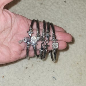 May include: A selection of silver bracelets with decorative charms. The bracelets have a twisted cable design and are embellished with charms including a key, a square of sparkling stones, and other decorative features.