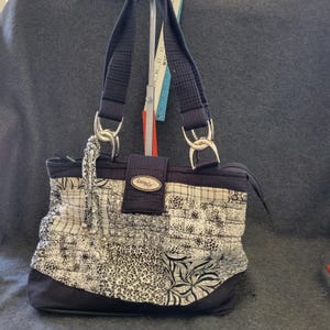 Puede incluir: Bolso tote con estampado en blanco y negro con herrajes plateados. El bolso presenta una variedad de estampados, incluyendo estampados florales y de animales. Las asas son negras y el bolso tiene un cierre de solapa con un pequeño logotipo.