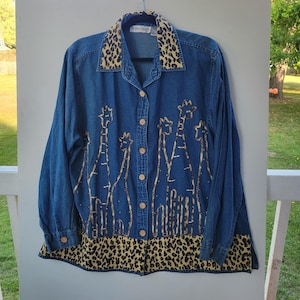 May include: Blue denim button-up shirt with long sleeves. The collar and hem feature a leopard print pattern. The shirt is decorated with giraffe silhouettes in a gold color.