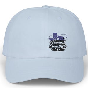 May include: Pale blue baseball cap with a curved peak. The cap displays a purple graphic of the Texas state outline, a fishing hook, and the words "Hooked Together TX" in black script.