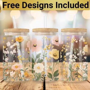 May include: Three clear glass tumblers with lids and straws, each featuring a different floral design with pink, yellow, and white flowers. The tumblers are sitting on a wooden surface.