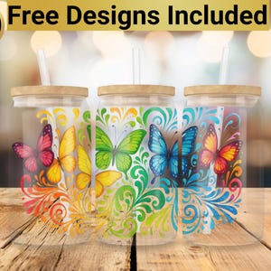 May include: Three clear glass tumblers with lids and straws, each featuring a different colorful butterfly design with floral accents. The tumblers are on a wooden surface.