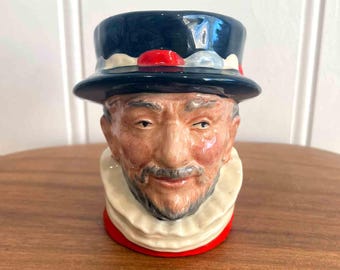 May include: A ceramic mug shaped like a man's head, featuring a black hat with red and white accents. The mug has a white ruffled collar and a red base. The face is detailed with a mustache and beard.