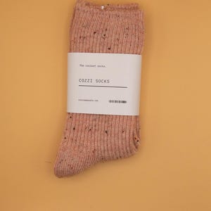 Puede incluir: Un par de calcetines rosados moteados con una etiqueta blanca que dice "The coziest socks. COZZI SOCKS cozzisocks.com".