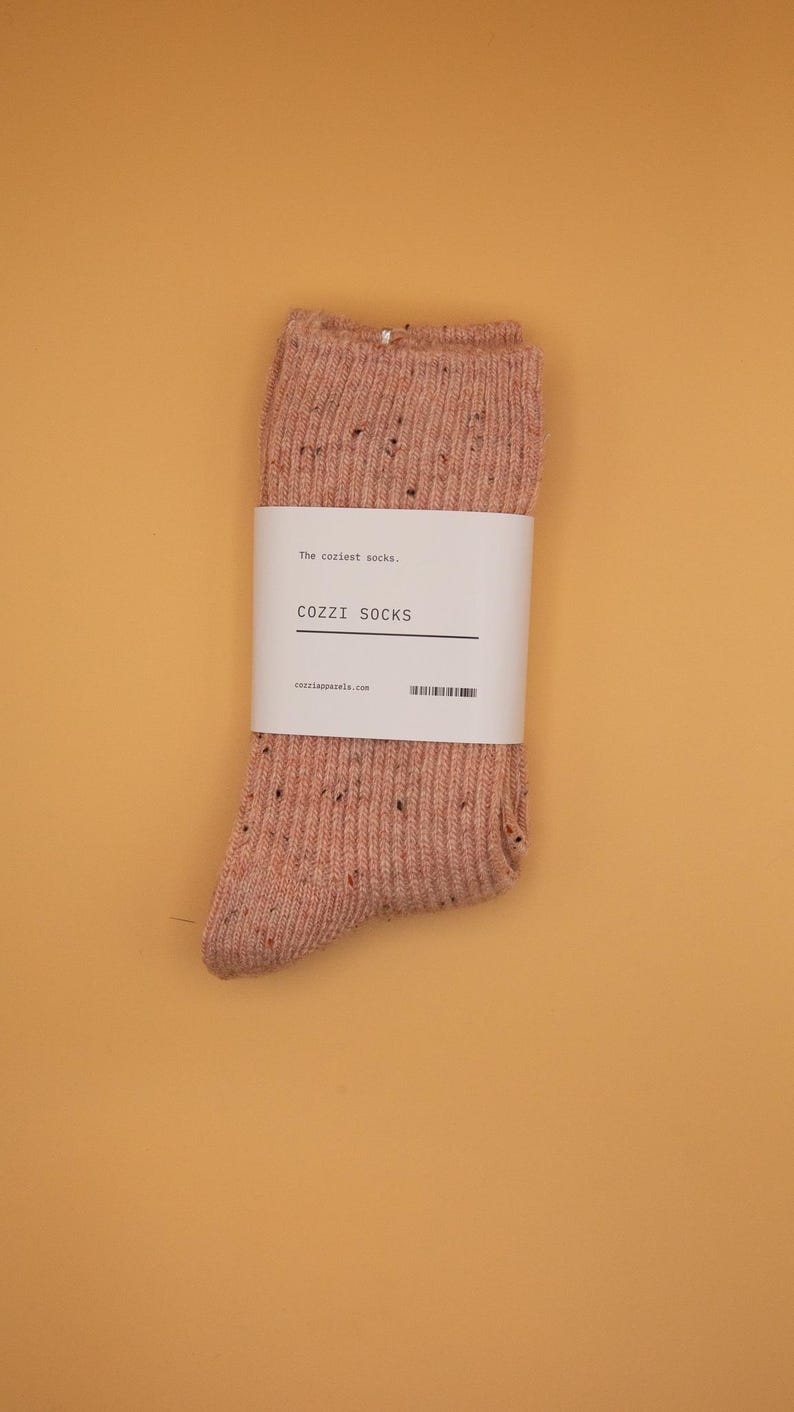 Puede incluir: Un par de calcetines rosados moteados con una etiqueta blanca que dice "The coziest socks. COZZI SOCKS cozzisocks.com".