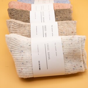 Puede incluir: Un conjunto de cinco pares de calcetines acogedores en varios colores, incluyendo beige, rosa claro y azul. Los calcetines est&aacute;n empaquetados con una etiqueta blanca que dice "The coziest socks. COZZI SOCKS cozziapparela.com".