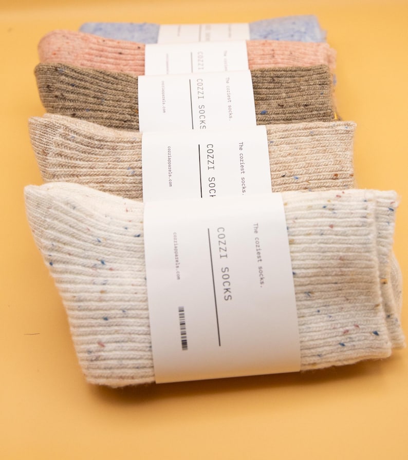 Puede incluir: Un conjunto de cinco pares de calcetines acogedores en varios colores, incluyendo beige, rosa claro y azul. Los calcetines est&aacute;n empaquetados con una etiqueta blanca que dice "The coziest socks. COZZI SOCKS cozziapparela.com".