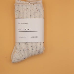 Puede incluir: Un par de calcetines blancos con un patr&oacute;n moteado. Los calcetines tienen una etiqueta que dice "The coziest socks... COZZZI SOCKS cozziapparel.com".