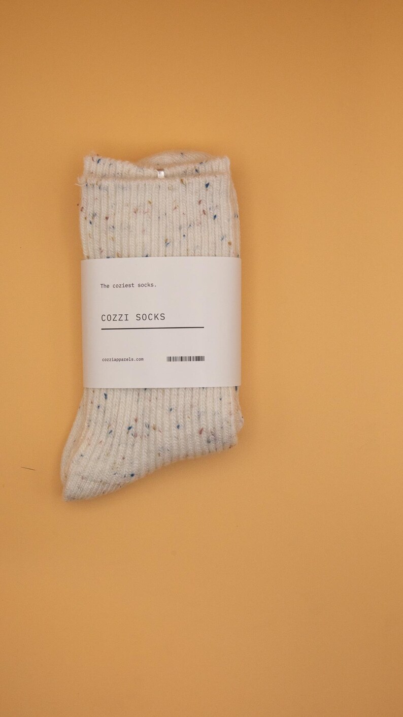 Puede incluir: Un par de calcetines blancos con un patr&oacute;n moteado. Los calcetines tienen una etiqueta que dice "The coziest socks... COZZZI SOCKS cozziapparel.com".