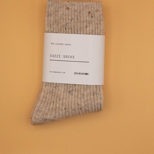 Puede incluir: Un par de calcetines beige con un patr&oacute;n moteado. Los calcetines tienen una etiqueta blanca que dice "The coziest socks." "COZZI SOCKS" y "cozzisocks.com".