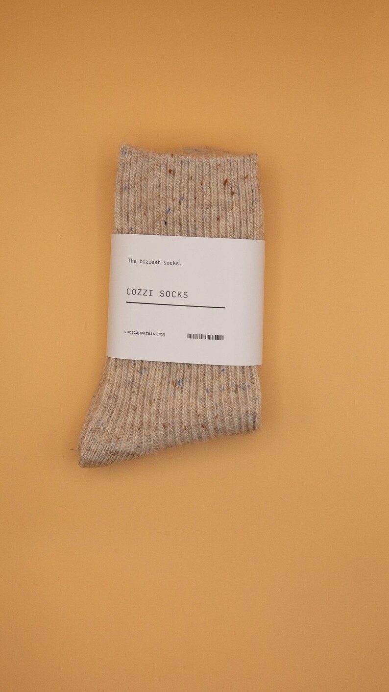 Puede incluir: Un par de calcetines beige con un patr&oacute;n moteado. Los calcetines tienen una etiqueta blanca que dice "The coziest socks." "COZZI SOCKS" y "cozzisocks.com".